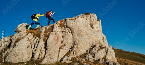 Couple hikers trek up a rocky mountain trail, enjoying a scenic view above the clouds, with majestic peaks in the background.