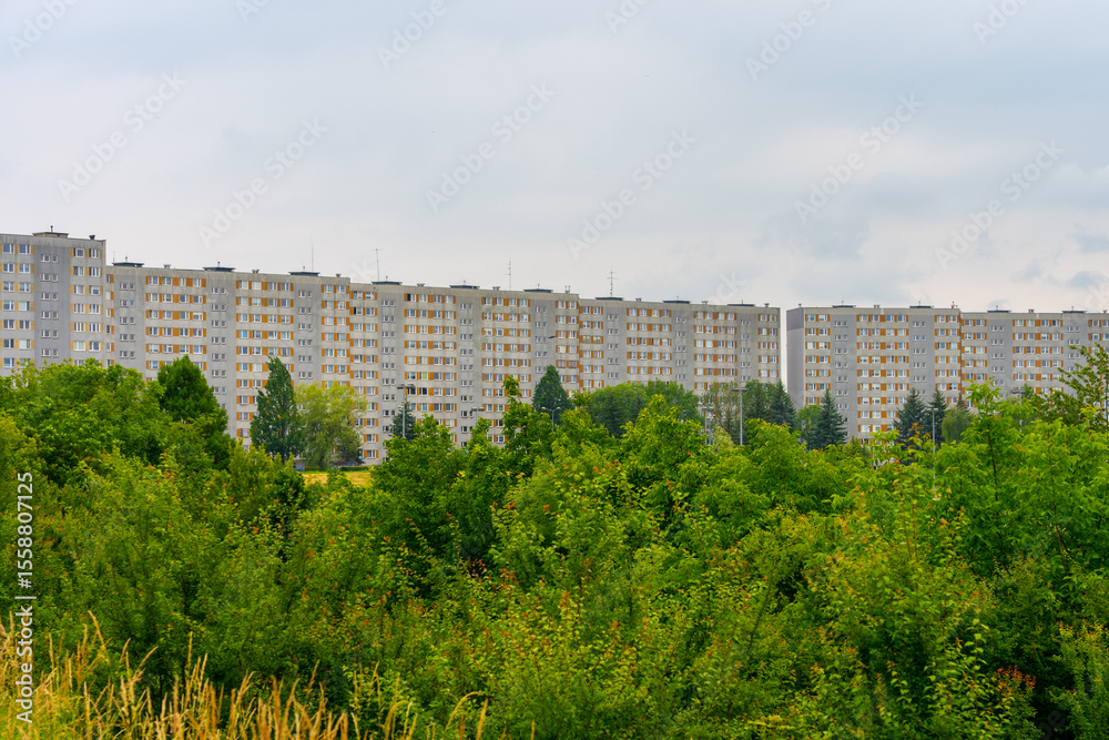 Naklejka premium Row of identical gray apartment blocks behind lush green trees under cloudy sky, post-communist housing estate concept
