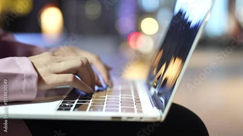 Close-up of a person typing on a laptop at night with blurred lights in the background, symbolizing technology, remote work, focus, and modern digital life.