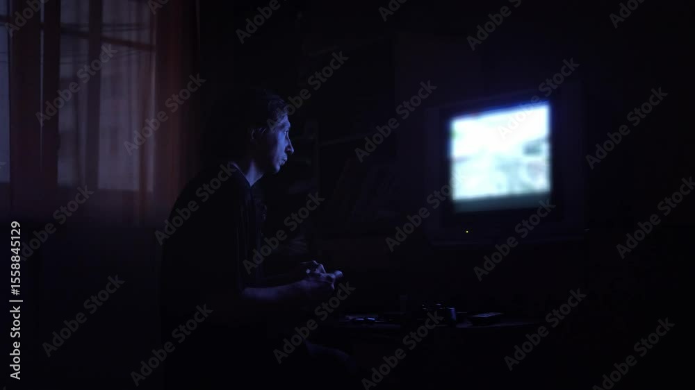 An intensely absorbed and atmospheric mood is captured as a man plays a video game in a dark room, his face illuminated by the blue glow of an older television screen