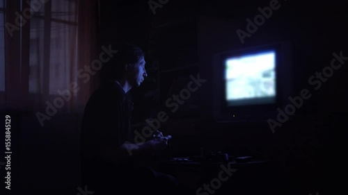 An intensely absorbed and atmospheric mood is captured as a man plays a video game in a dark room, his face illuminated by the blue glow of an older television screen