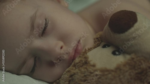 the little kid sleeps hugging a teddy bear while lying in bed, boy dreams with soft toy, covered with warm blanket in children's room, closed eyes of child rest at night, doze in dark with toy friend.