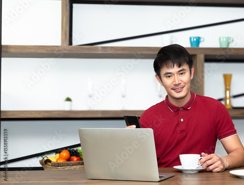 Young Smiling handsome wearing red t-shirt meeting online and drinking a cup of coffee and
work from home. Communicating online, writing emails, distantly working or studying on computer.