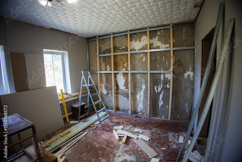 Metal frame attached to the wall with wooden bars and screws, preparing for plasterboard installation in a renovated room with scattered construction materials and a nearby ladder.