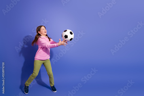 Cheerful schoolgirl in casual fashion catching a soccer ball on a purple background, symbolizing active childhood joy