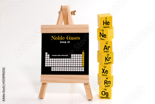 Six yellow cubes, each labeled with the symbols and atomic numbers of noble gases, arranged beside a chalkboard displaying the Periodic Table of Elements, with Group 18 highlighted in yellow