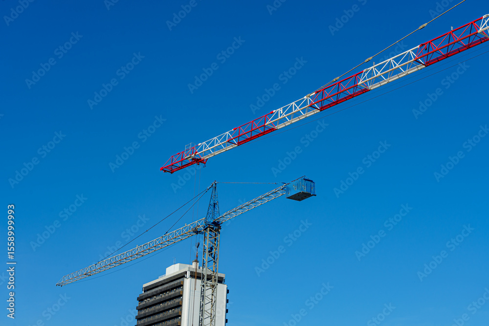 Sticker Two large construction cranes, one red and white, the other blue, dominate the skyline above a building, set against a vibrant blue sky
