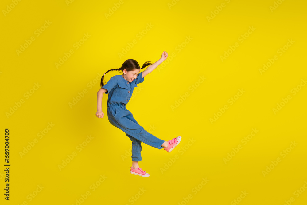 Obraz premium Preteen girl joyfully jumping in a stylish blue outfit against a vibrant yellow background, expressing happiness