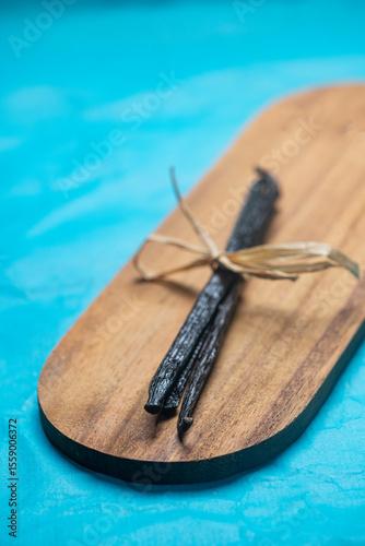 Close up studio still life photo featuring dried vanilla beans on a vintage wooden board and vibrant blue hand made paper background , ideal for culinary, organic and gournet food concepts