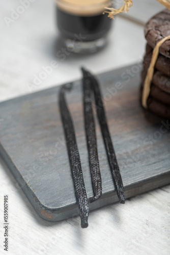 Close up studio still life photo with soft light and muted colors featuring dried vanilla beans on a vintage wooden board  with espresso and cookies in the background