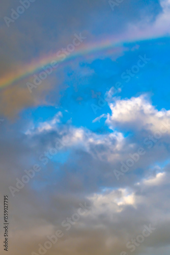 Rainbow in cloudy blue sky. Natural weather phenomenon with scattered clouds. Atmospheric landscape and nature background. Design for poster, wallpaper, postcard, greeting card. Vertical outdoor shot.