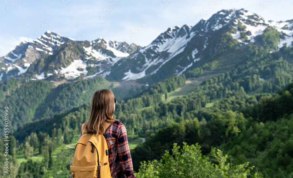 Naklejka premium Young woman with a yellow backpack stands on a mountain trail, gazing at the majestic snow-capped peaks and lush green valleys, embodying adventure and exploration in nature, copy space
