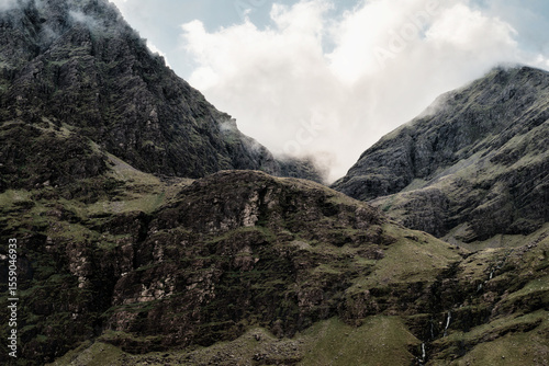 Fog drifts over rocky, green terrain leading up to the peak of Carrauntoohill in County Kerry, Ireland. A small waterfall trickles down the rugged mountain face.
