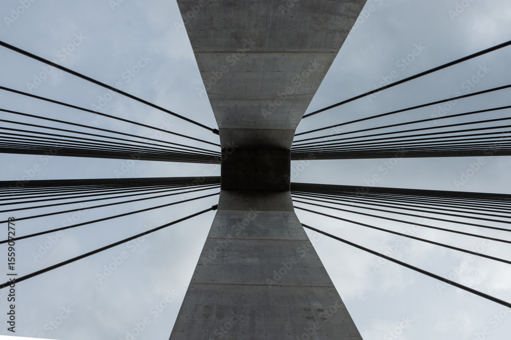 Fototapeta premium 埼玉県秩父市の合角漣大橋の橋脚を真下から見上げた、抽象的な建築写真。コンクリートの主塔から放射状に伸びるケーブルが作り出す、幾何学的で力強いデザイン。近代的な土木技術やインフラ、アートを象徴するイメージ。