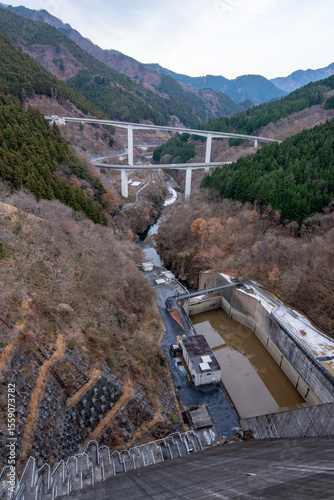 埼玉県秩父市の滝沢ダムの堤体から見下ろした、冬の渓谷の風景。眼下にはダムの放流設備、遠くには巨大なループ橋「雷電廿六木橋」を望む。日本の壮大なインフラと、厳しい冬の自然が織りなす絶景。

