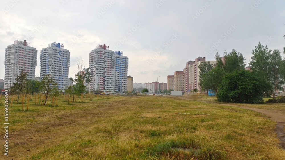 Trees grow on a lawn with a walking path and sun-baked grass. Behind them is a city street with buildings, traffic lights and parked cars. Cloudy summer weather and it's raining Trees grow on a lawn with a walking path and sun-baked grass. Behind them is a city street with buildings, traffic lights and parked cars. Cloudy summer weather and it's raining