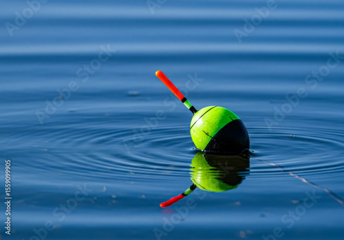 A bright green and black fishing float with a red top casts a reflection on the blue water surface, creating gentle ripples.