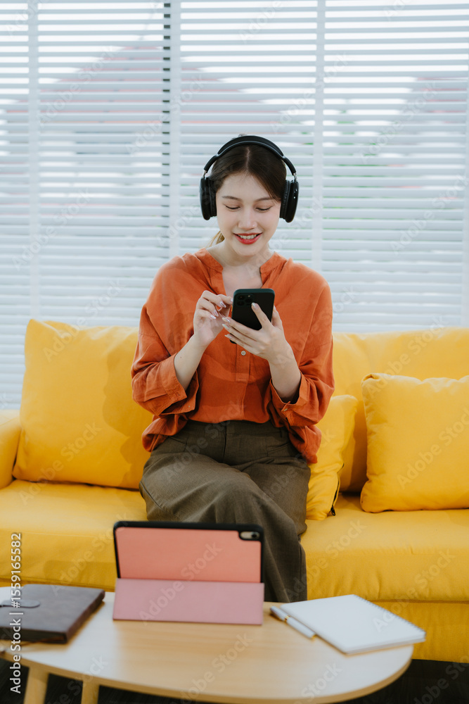 Fototapeta premium Young asian woman using smartphone and wearing headphone sitting on couch at home. Happy girl relaxing comfortable in the living room, spending lazy weekend, listening to music, watching movie.