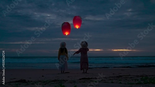 Two young women release lit paper lanterns at dusk on the beach. Two friends release paper lanterns into the sky