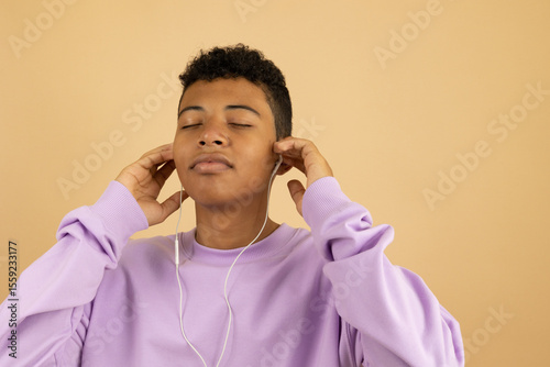 Brown skinned Latin American teenager wearing a lilac sweatshirt with his eyes closed feeling the rhythm, listening to music with wired headphones on a light brown background with copyspace