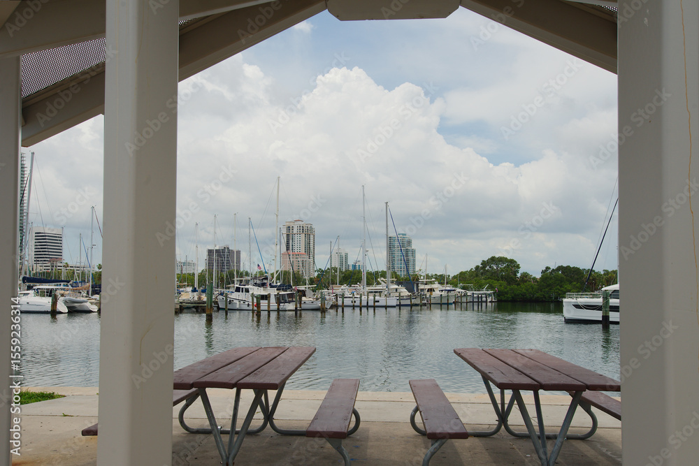 Fototapeta premium City Skyline and Waterfront with Boats Under Bright Sky St. Petersburg, FL. Beautiful cityscape featuring tall buildings near a waterfront with boats docked. Framed park bench. 