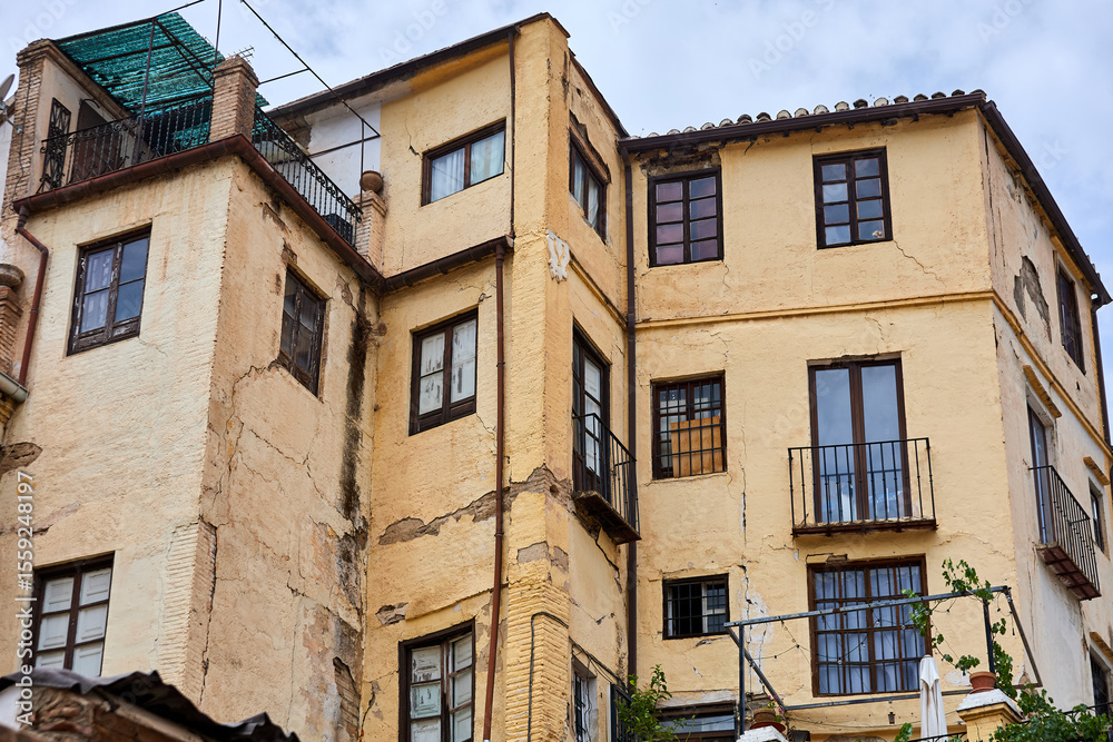 Obraz premium Old building with yellow stucco walls, cracked surfaces, wooden windows, small balconies, and tiled roof under overcast sky; architecture, facade;