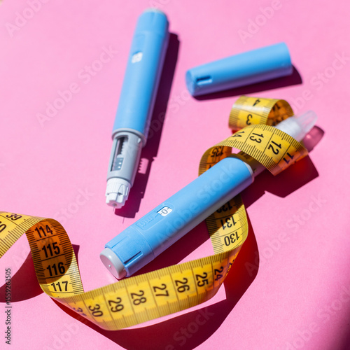 Blue Ozempic pens and tape on bright pink table. Abstract representation of health, wellness, and medical aid. Yellow measuring tape around the injectors.