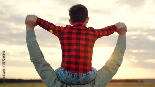 Father holds boy on shoulders spreading arms to sides at sunset on field with breathtaking view of nature beauty. Father spends time with little boy. Calm boy feels warmth and care from father