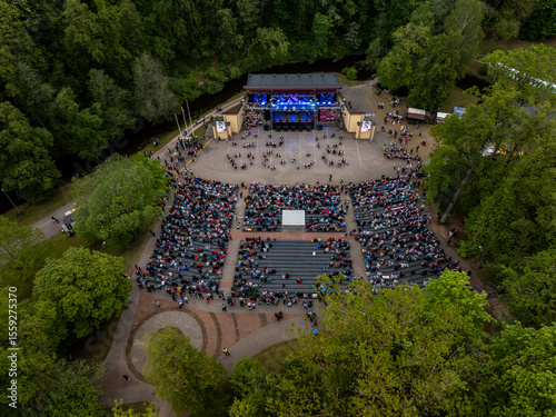 An aerial view of an outdoor amphitheater surrounded by trees, featuring a stage with blue lighting, seated audience, and distinctive circular pathways.