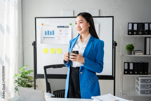 A woman in a blue jacket is holding a cup of coffee in front of a white board wi