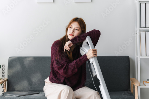 Exhausted Asian Woman with Vacuum Cleaner Sitting on Sofa Feeling Tired of Cleaning Housework Looking Away Thinking About Resting After Cleaning