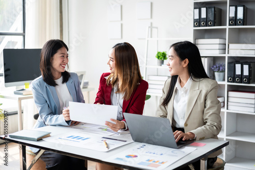 Three women are sitting at a desk with papers and laptops