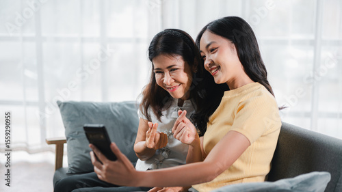 Asian Adult Woman and Older Asian Woman Taking Selfie Together on Mobile Phone on Sofa in Living Room Smiling and Posing Mini Heart Hand Sign At Home