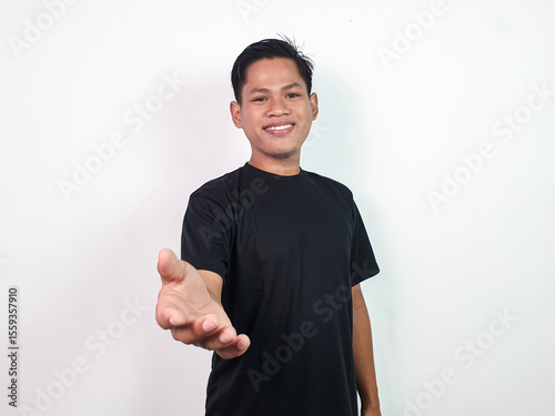 Bild auf Leinwand Young Asian Man in Black Shirt Smiling Extending Hand Forward in Friendly and Welcoming Gesture, Standing Against Plain White Background
