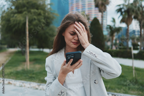 Woman stressed by financial crisis reading stock market fall news on smartphone in urban outdoor setting with green palms and buildings
