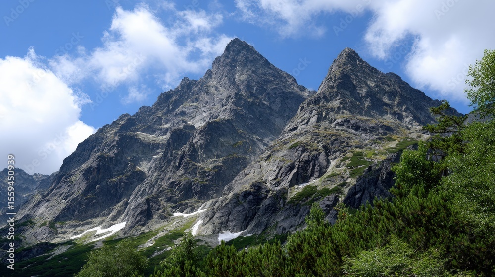 Obraz premium Majestic mountain peaks under a clear blue sky with scattered clouds, surrounded by lush green foliage and patches of snow.