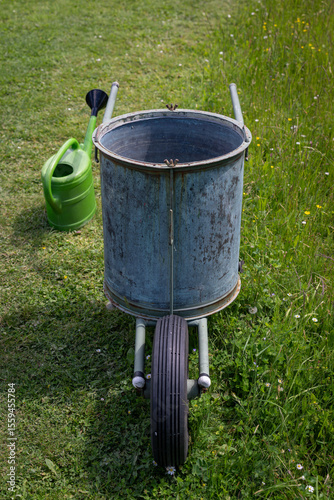 Wallpaper Mural Metal water container on a lawn with partially mown grass and a green plastic watering can, captured in natural outdoor light, highlighting the contrast between utilitarian objects and garden textures Torontodigital.ca