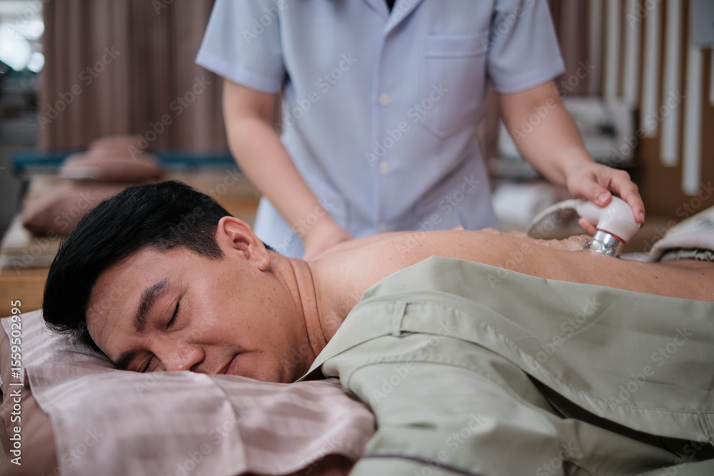 Obraz premium Male Asian patient recovery treated by gel ultrasound device for back pain relief procedure by female physical muscle doctor, passive medical therapy at spa clinic, and wellness massaging healthcare.