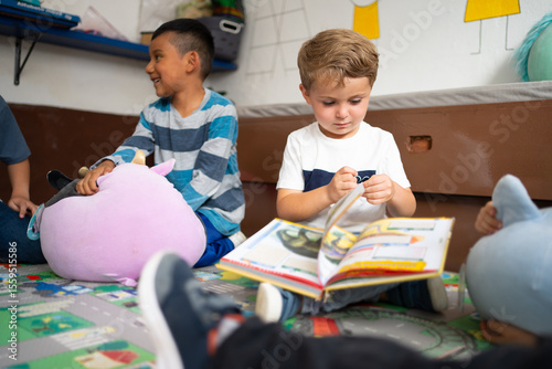 A kindergarten boy sits on the classroom floor looking closely at a storybook during preschool hours. Concept of early childhood literacy, imagination, and focus through reading activities