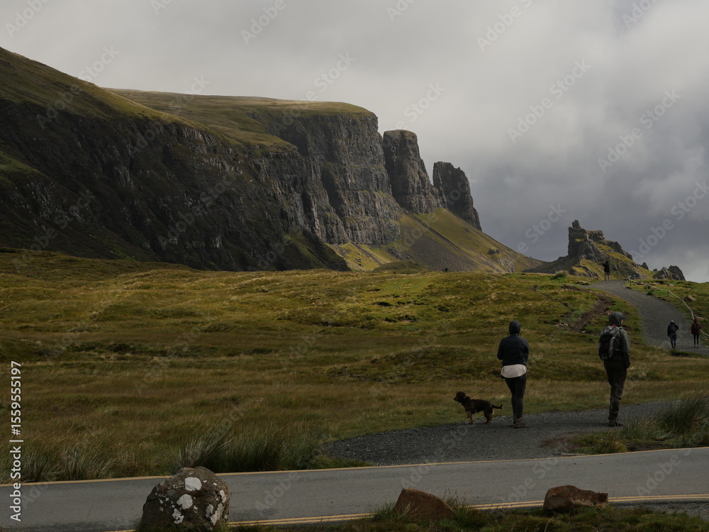 Fototapeta premium Hikers on a scenic trail at the Quiraing, Isle of Skye, Scotland, surrounded by dramatic cliffs and lush green landscape under a cloudy sky. Outdoor adventure and nature travel.