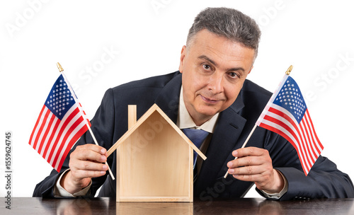 Middle aged man in business suit holding american flags in hands and miniature symbolic wooden house on white background