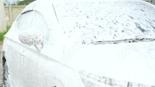 A middle-aged Asian man carefully washes his car using water and foam. Each step from high-pressure rinsing to scrubbing and drying ensures the vehicle is spotless, well-maintained, and presentable