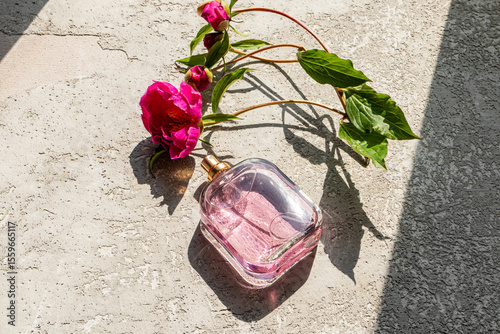 A transparent bottle of cosmetic spray or women's perfume lies on a gray cement background with pink peony buds. A blank bottle mockup. Sun shadows.