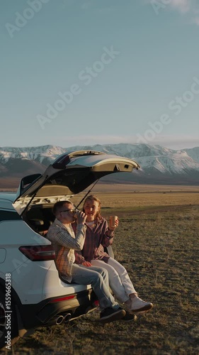A young couple of tourists in love, a man and a woman, are sitting in the trunk of a car, drinking coffee and admiring the sunset and mountain views. Vertical video.