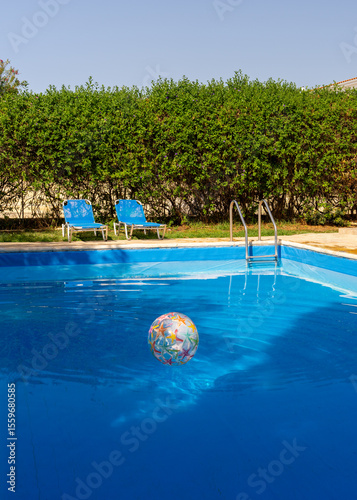 Bright inflatable beach ball drifting in swimming pool next to two empty lounge chairs. Playful and sunny vacation vibe, perfect for family, fun, and leisure content.