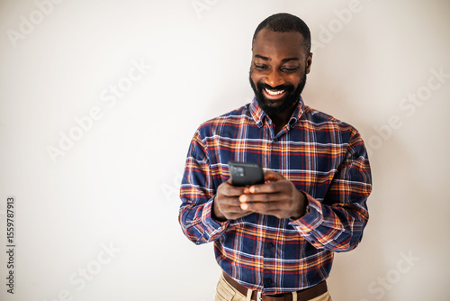 Happy black man in cozy shirt shirt standing in front of wall and messaging on smartphone.