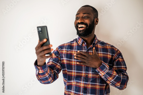 Ecstatic black man in cozy shirt standing in front of wall and having video call on smartphone.