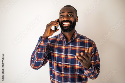 Cheerful black man in cozy shirt standing in front of wall and talking on mobile phone.