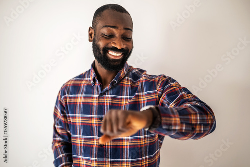 Cheerful black man in cozy shirt standing in front of wall and looking at his wristwatch.