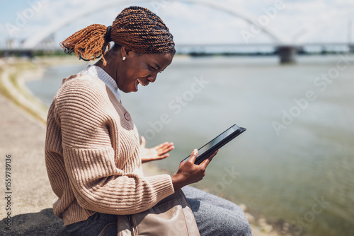Young stressed black woman is using digital tablet. She is sitting by the river in the city.